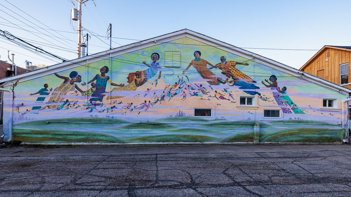 A mural on a buildings end depicting colorfully dressed figures dancing and running across a pastel landscape.