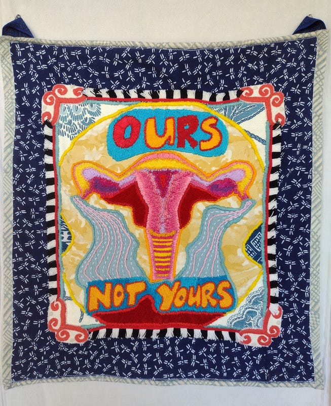 A colorful embroidered uterus in reds, pinks, yellows, and lilac with Ours Not Yours embroidered above and below it in golden yellow, orange, red, and turquoise. 