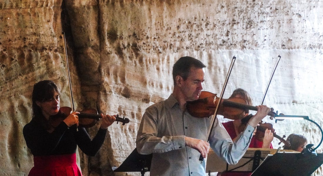 Two women and one male playing string instruments, with a cave in the background Two women and one male playing string instruments, with a cave in the background