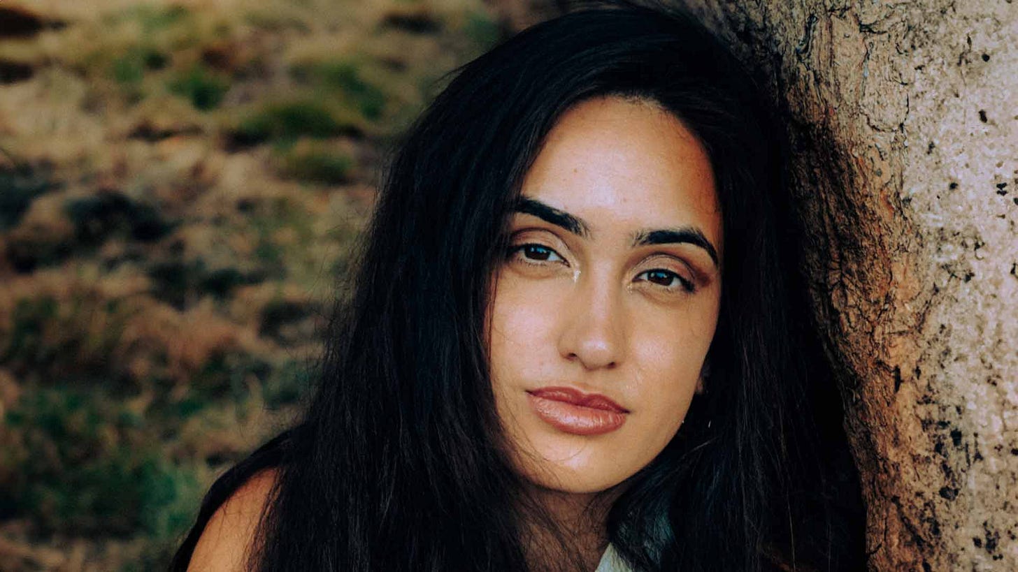 An image of Tice Cin, a British Turkish Cypriot woman with long dark hair and glittery eye makeup. She is sitting outside leaning against a tree.