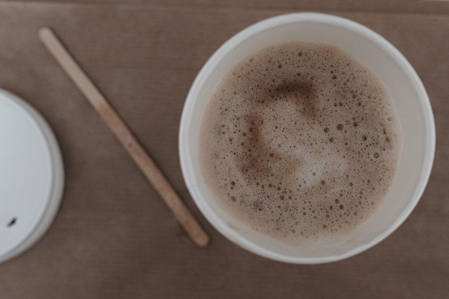A paper cup of coffee next to a wooden stirrer and a white plastic lid, set against a soft beige tabletop capturing a quiet café moment.