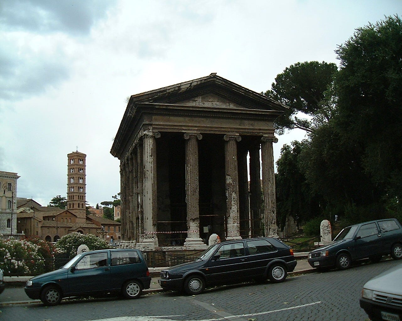 Three small dark-coloured cars parked on a slop in front of a small but traditional temple that has foru columns across the front. In the background the tall bell tower of Santa Maria in Cosmedin in the background