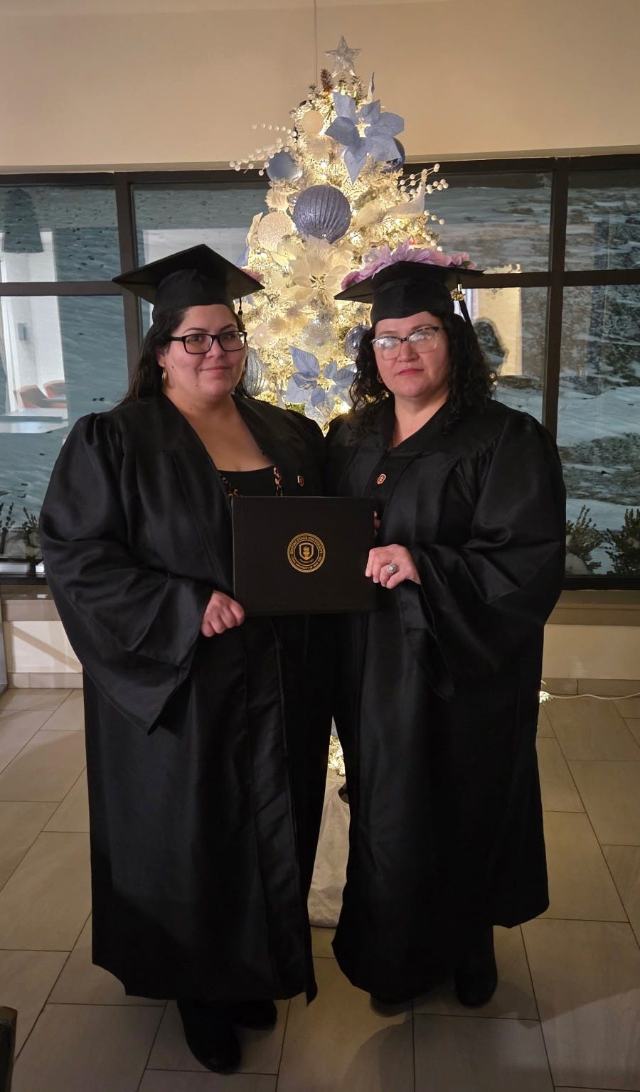 Two women, both wearing black graduation gowns and caps, stand in front of a Christmas tree. They are holding up a diploma.