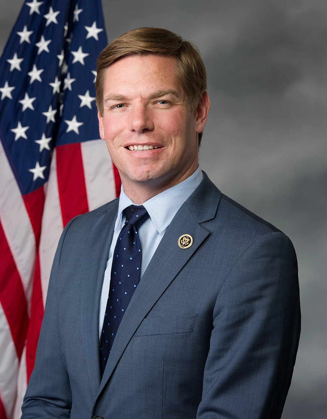 Swalwell official portrait; in front of flag. he is middle aged white man; brown hair, wearing dark suit and tie. smiling over forcefully