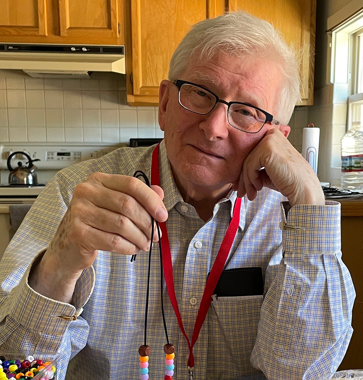 An older man with white hair and glasses, wearing a light plaid shirt with pale blue and yellow lines and a red lanyard, sits at a kitchen table with a gentle smile. He rests his head on one hand and holds up a beaded necklace made at Yunasa. An older man with white hair and glasses, wearing a light plaid shirt with pale blue and yellow lines and a red lanyard, sits at a kitchen table with a gentle smile. He rests his head on one hand and holds up a beaded necklace made at Yunasa.