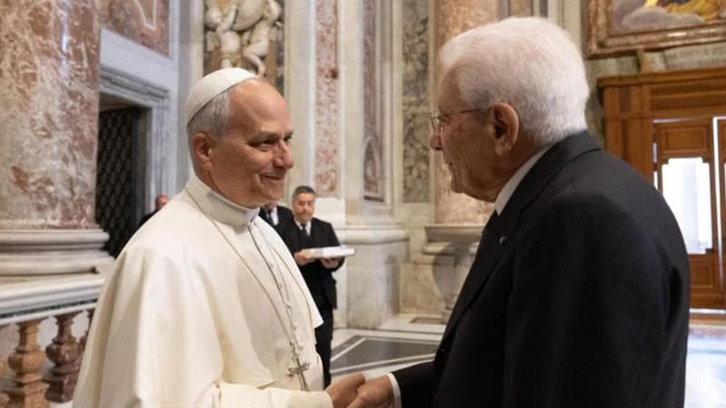 Papa in abito bianco (zucchetto e croce pettorale) stringe la mano al presidente della Repubblica italiana Sergio Mattarella in abito scuro, all’interno di una sala riccamente decorata con marmi e affreschi; sullo sfondo una terza persona osserva la scena.
