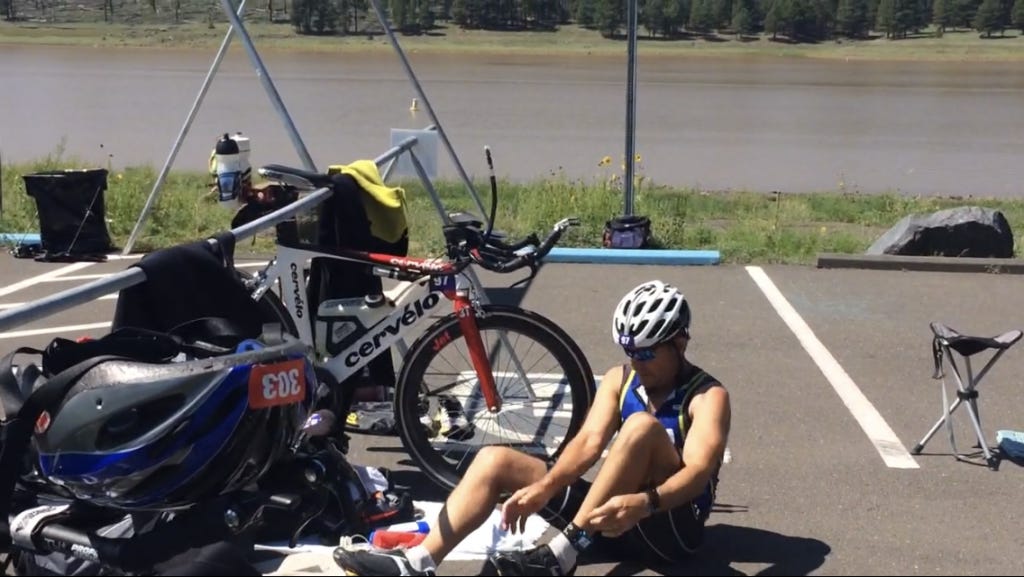 Athlete sitting on the ground changing gear during triathlon transition next to a rack of triathlon bikes.