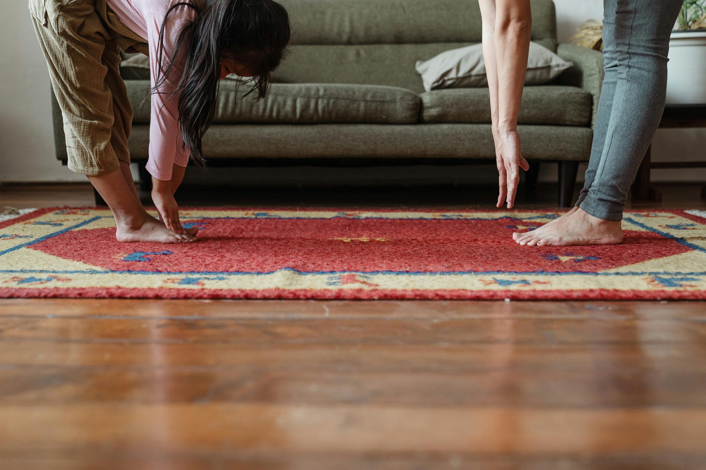 A low-angle photo shows a woman and a child stretching. Both are barefoot and are bending over from the waist, reaching for their feet. They are standing on a patterned red and yellow rug, in a room with a green sofa in the background.