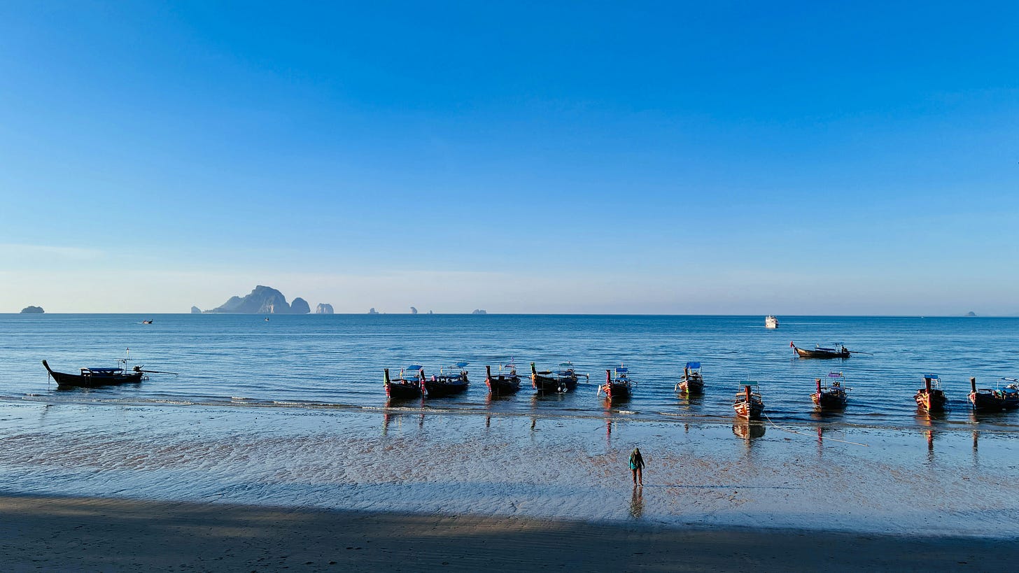 A wide landscape photograph titled "Phuket Coast.webp" showing a line of traditional wooden longtail boats anchored in the shallow water along a sandy beach under a clear blue sky in Phuket, Thailand. A lone figure stands near the shoreline, looking out toward the calm turquoise sea. In the far distance, distinct limestone cliffs rise out of the ocean horizon. The image serves as a visual backdrop for the newsletter's focus on Southeast Asia's vast coastlines, marine ecosystems, and the growing regional push toward sustainable maritime policies to combat climate change. A wide landscape photograph titled "Phuket Coast.webp" showing a line of traditional wooden longtail boats anchored in the shallow water along a sandy beach under a clear blue sky in Phuket, Thailand. A lone figure stands near the shoreline, looking out toward the calm turquoise sea. In the far distance, distinct limestone cliffs rise out of the ocean horizon. The image serves as a visual backdrop for the newsletter's focus on Southeast Asia's vast coastlines, marine ecosystems, and the growing regional push toward sustainable maritime policies to combat climate change.