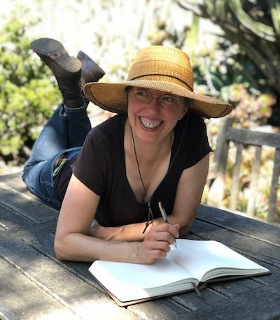 Grace lying on stomach on park table outdoors, up on elbows writing in journal, smiling, wearing sun hat, jeans, boots