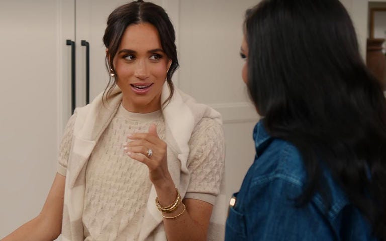A photo of Meghan Markle in a beige kitchen wearing a beige sweater with another beige sweater wrapped around her shoulders. It's just beige all the way down.