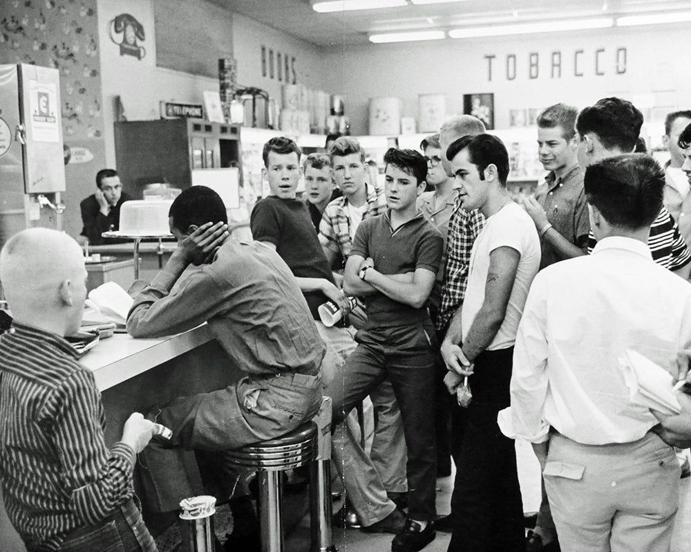 Black and white photo of Dion Diamond staging a sit-in at a drugstore counter surrounded by an angry crowd of white people