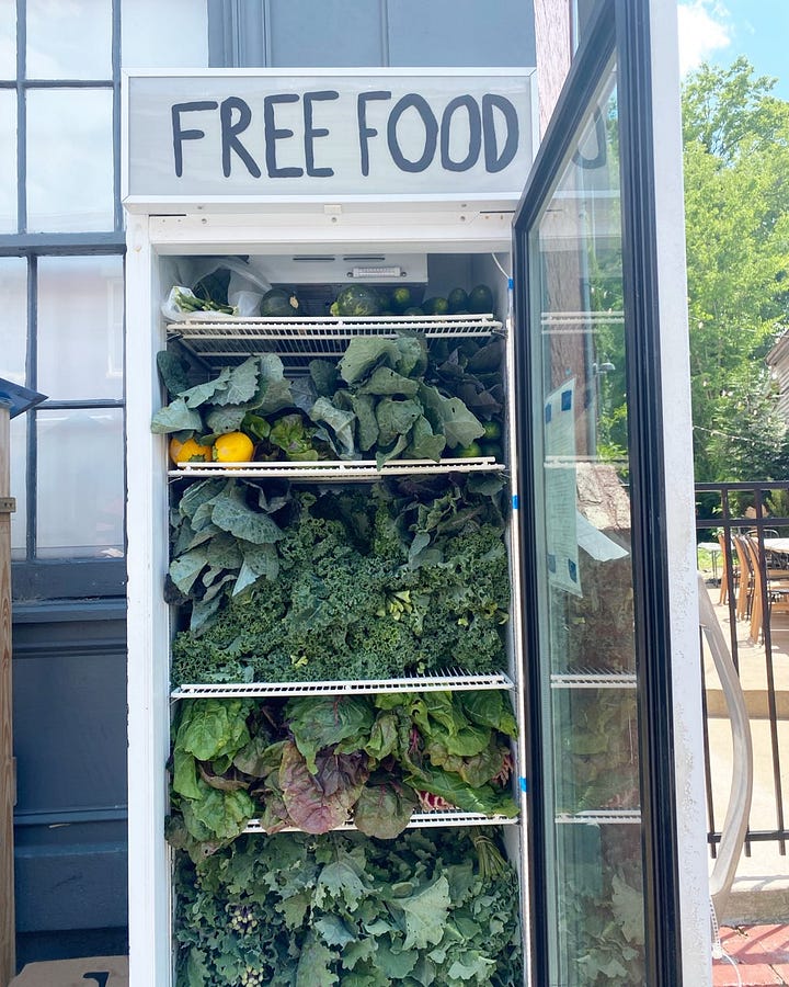 A fridge outside a cement building with lots of colorful flowers painted on it and the words 'FREE FOOD' on it, and a fridge open and full of tons of fresh produce.