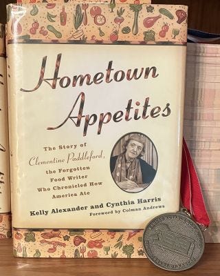 The book cover of Hometown Appetites by Kelly Alexander and Cynthia Harris. It is has photo of Clementine Paddleford. Beside the book is a metal that accompanied the Kansas Notable Book Award.