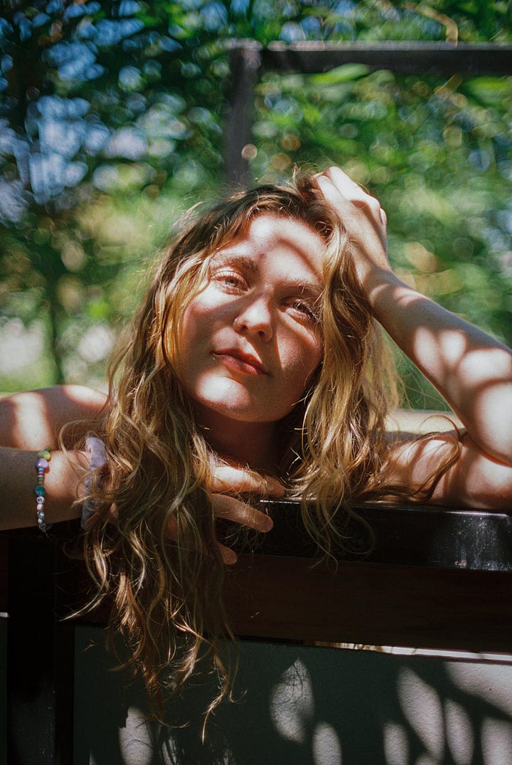 First image is a person with blonde hair surrounded by greenery and shadows. Second image is a group of four people standing together. Third image is of a person wearing glasses and playing guitar on a stage. Fourth image is of five people hugging in front of a wooded area. 