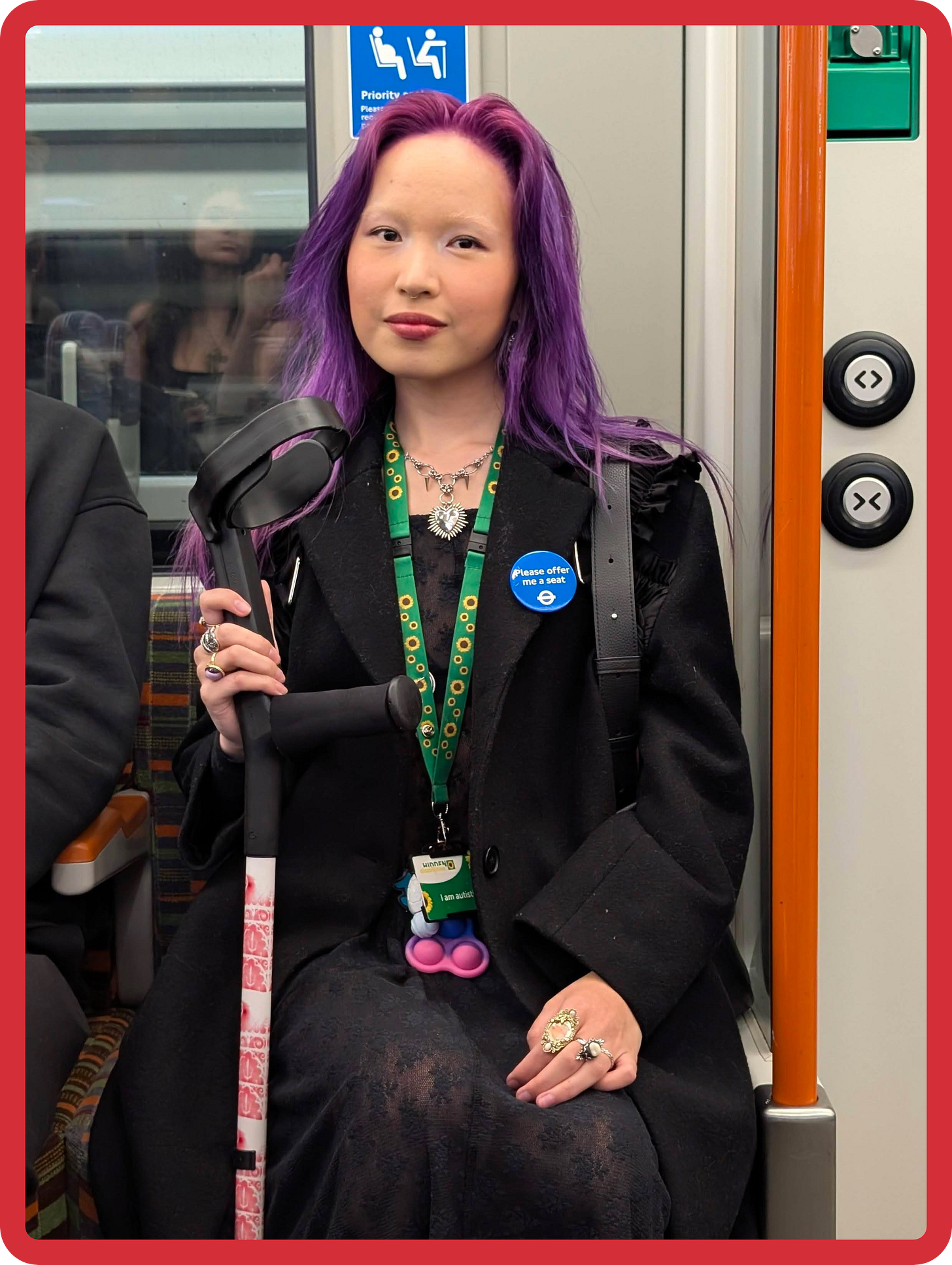 A young person who is dressed in black and has long purple hair, sitting down in a priority seat on a London Overground train. They are wearing a green sunflower lanyard around their neck, and a blue TfL 'Please offer me a seat' badge on their coat. They are also holding a crutch in one hand, and smiling gently at the camera. A young person who is dressed in black and has long purple hair, sitting down in a priority seat on a London Overground train. They are wearing a green sunflower lanyard around their neck, and a blue TfL 'Please offer me a seat' badge on their coat. They are also holding a crutch in one hand, and smiling gently at the camera.