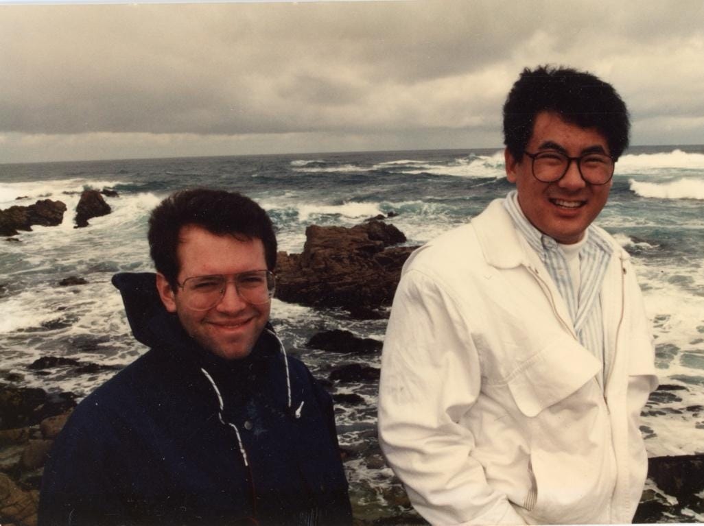 Elliot Schwartz (left) and Stephen Pao (right) smiling on the rocky California coast in the early 1990s, with waves crashing behind them. Elliot Schwartz (left) and Stephen Pao (right) smiling on the rocky California coast in the early 1990s, with waves crashing behind them.