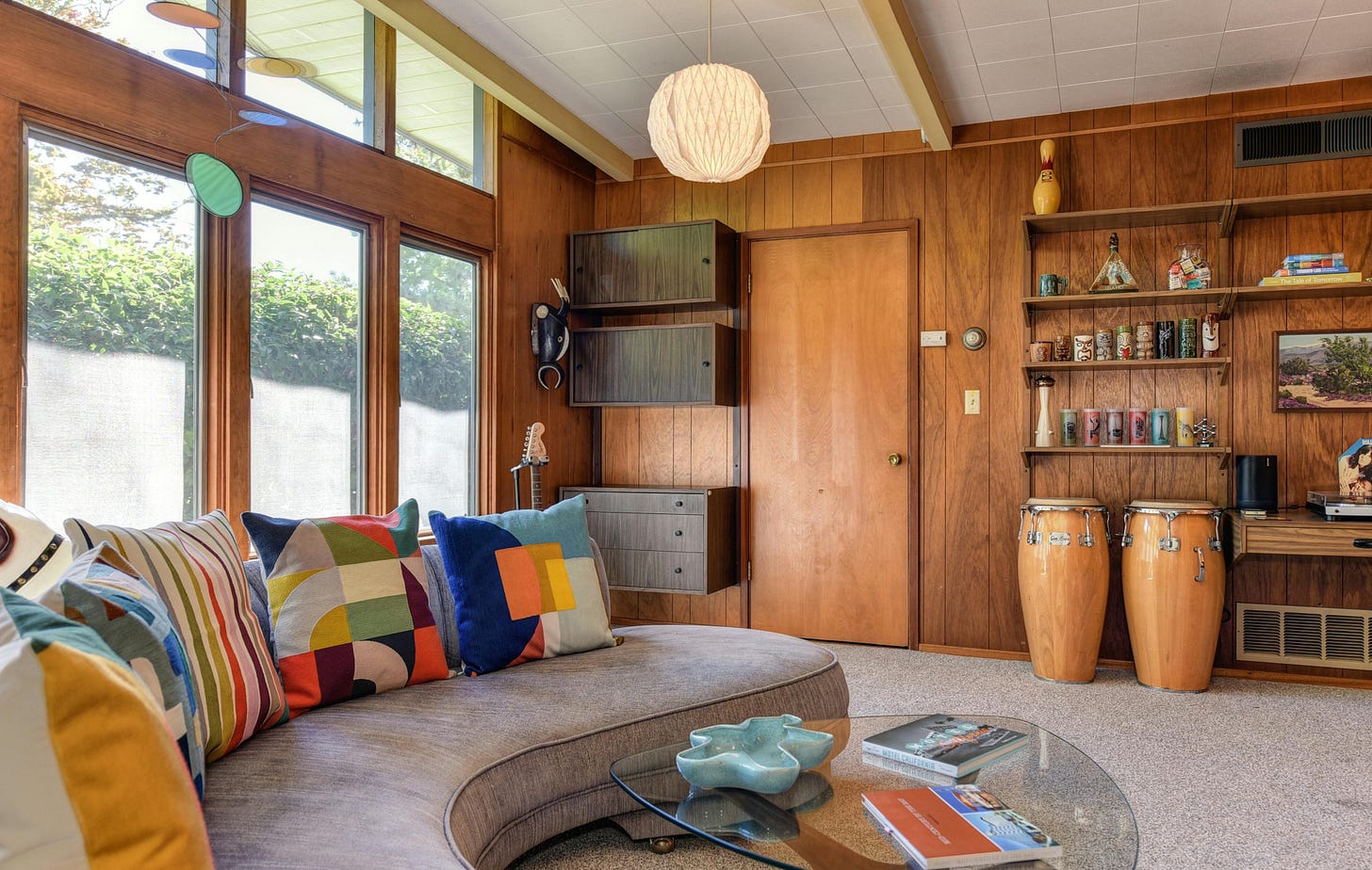 Wood-paneled room with a grey couch covered with multi-patterned pillows. A pair of conga drums sit by the door. A mid-century globe light fixture hangs from the ceiling Wood-paneled room with a grey couch covered with multi-patterned pillows. A pair of conga drums sit by the door. A mid-century globe light fixture hangs from the ceiling