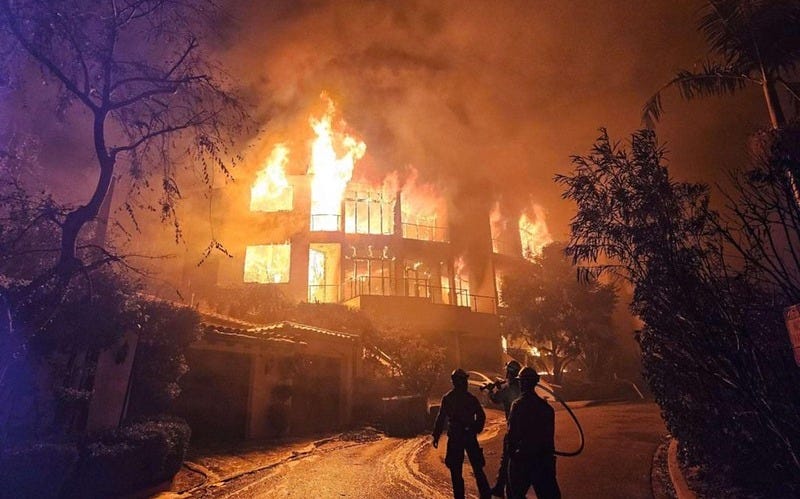 Firefighters aim hoses on the vegetation surrounding an unburned house (or outbuilding) on a hilly street, just below a larger two-story luxury home next door that is fully engulfed in flames. Orange flames light the scene, and the three firefighters silhouetted in the foreground appear tiny compared to the blaze.