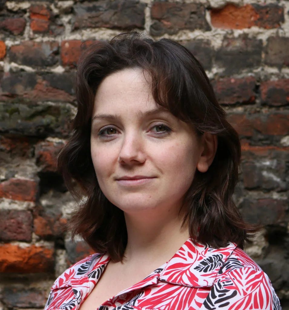 Polly is a white woman with dark brown hair. In this photo, she's standing in front of a brick wall wearing a red and black patterned blouse. Her hair is down and grazing her shoulders. She's turned towards the camera, with a gentle, closed smile.