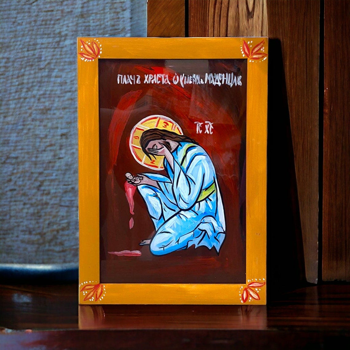 May include: A framed painting of a religious icon with a yellow frame and a red, orange, and blue background. The icon depicts a person kneeling with their head bowed and their hands clasped in prayer. The text on the painting reads 'Плач Христа о грешном Магдалине' and 'IC XC'. 
