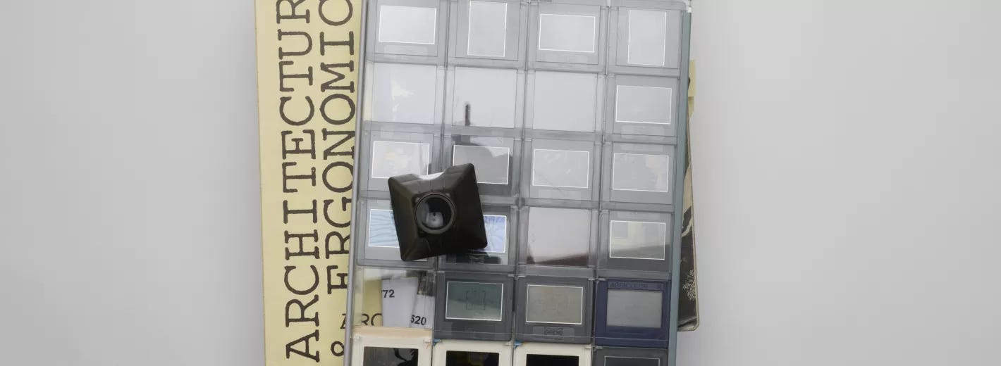 A stack of archival material rests on a light surface, topped with a clear sheet of photo slides arranged in a grid. A small black slide viewer rests on top, and sticky notes stick out from the pages.