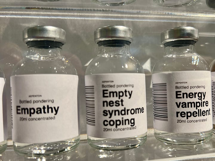 Art installation titled 'Bottled Pondering' by Aepenton, on display at the Design Museum Denmark. The piece features a wall-mounted grid of small glass vials, each labeled with a different abstract or emotional concept, such as 'Inner Critic Softener' or 'Courage Enhancer.' The vials are arranged in neat rows within a transparent display case, evoking the look of a scientific archive or apothecary cabinet of thoughts and feelings.
