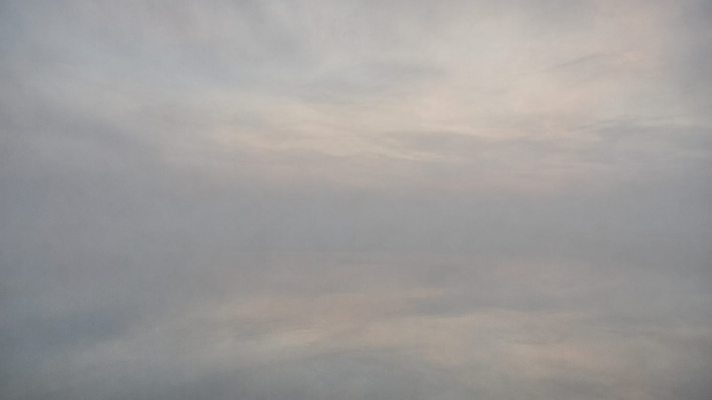 A calm river at first light reflecting a luminous sky. The boundary between water and atmosphere is nearly invisible, creating a contemplative sense of uncertainty about what is reflected and what is real.