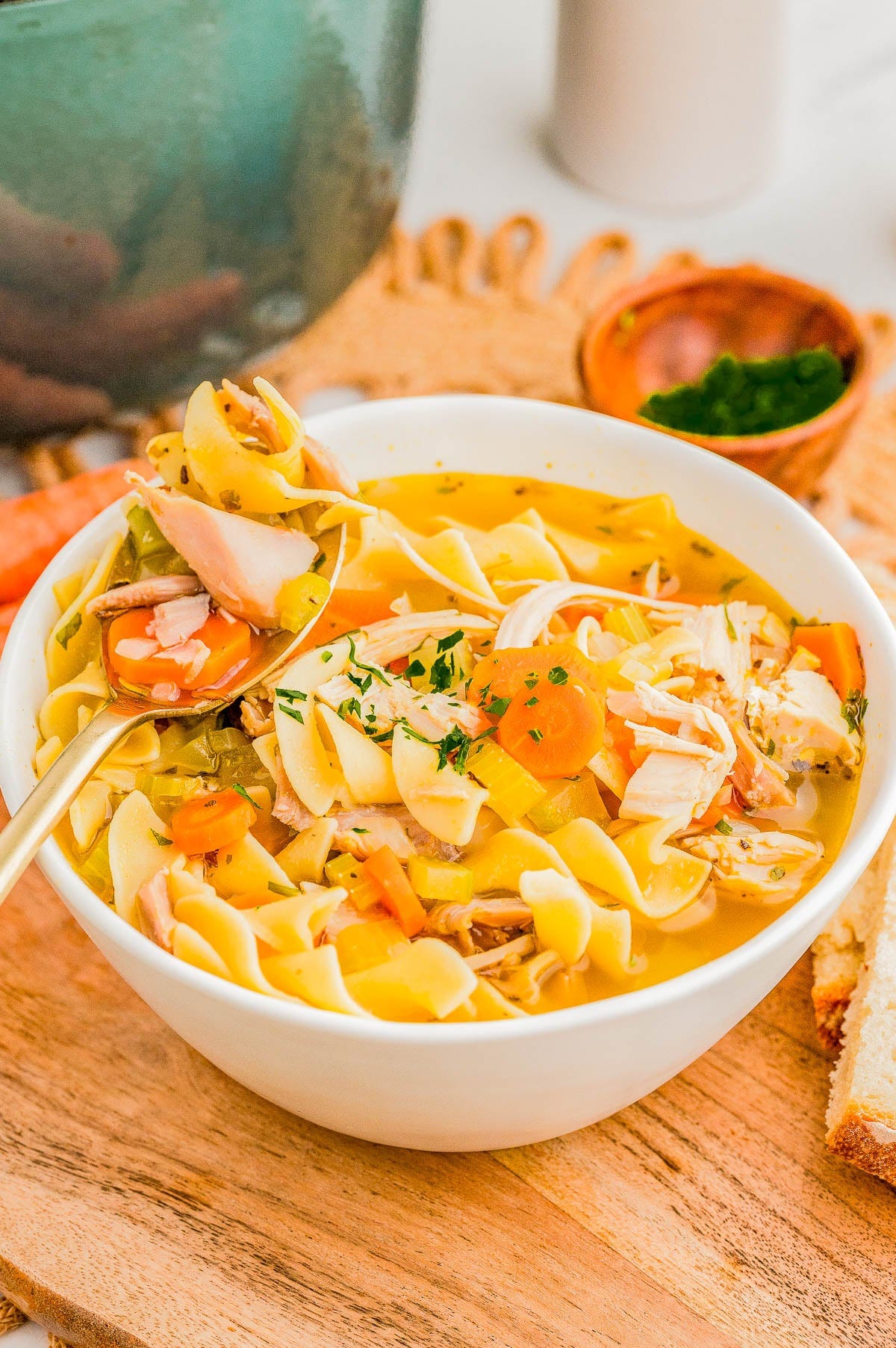 A bowl of chicken noodle soup with vegetables and herbs on a wooden board, accompanied by slices of bread and a small bowl of chopped parsley. A bowl of chicken noodle soup with vegetables and herbs on a wooden board, accompanied by slices of bread and a small bowl of chopped parsley.