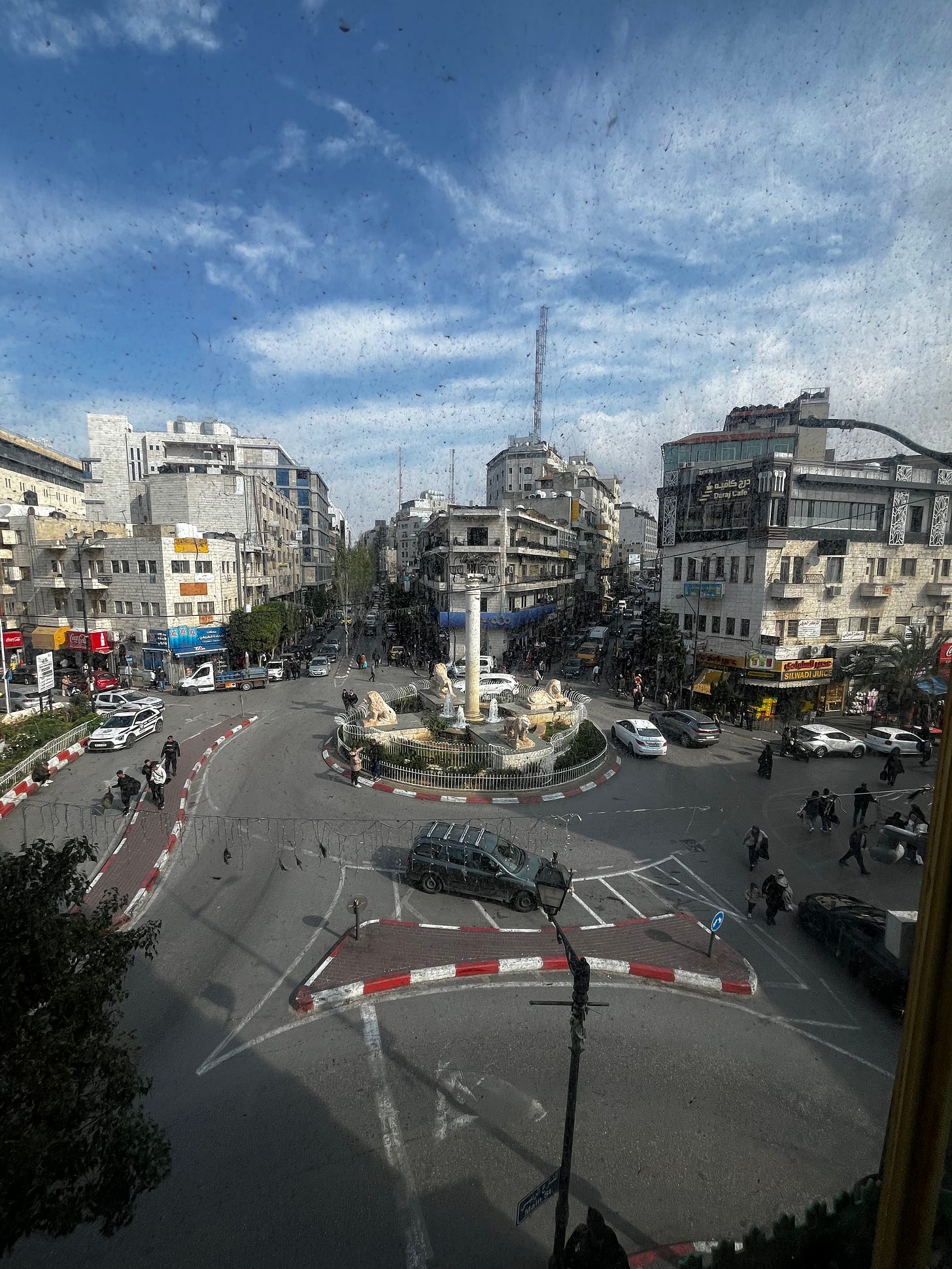 Elevated view through a rain-spotted window looking down on a busy urban roundabout. A stone column topped with a lamp and surrounded by four lion sculptures sits at the center of the traffic circle. Multi-story stone and concrete commercial buildings line the surrounding streets; pedestrians and cars move through the intersection beneath a partly cloudy sky.