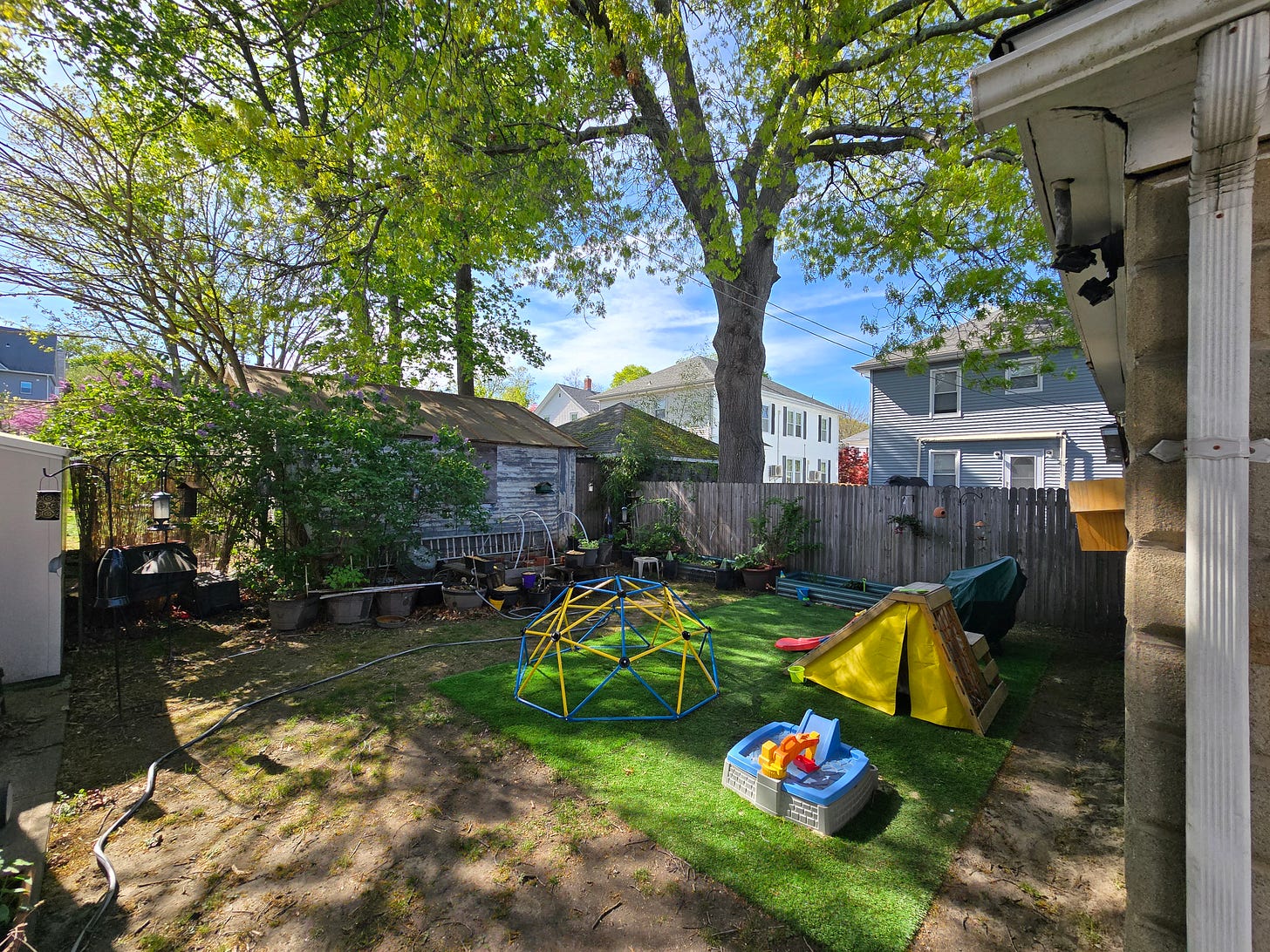 A wide shot of a small urban backyard, with several pieces of kids' play equipment on the right and containers and raised beds on the left.