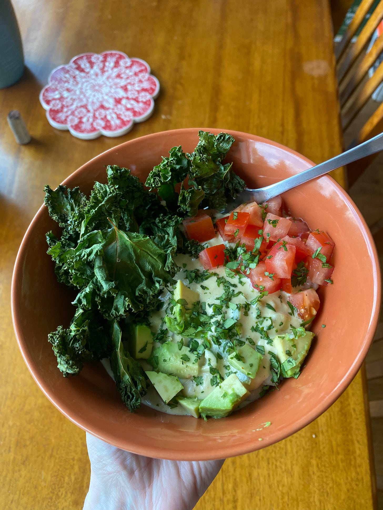An orange bowl, held in my hand, with the queso bowl described above. You can't see much of the rice and beans through the creamy sauce. Arranged on top of the sauce are chopped tomatoes and avocado, sprinkled with cilantro, and there are lots of kale chips piled on one side. In the background is the dining table with a red patterned trivet on it.