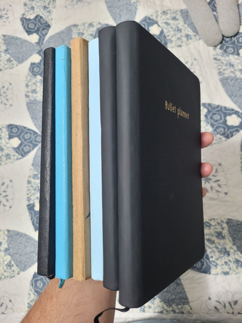 Minha mão segurando 6 cadernos fechados. A imagem permite ver as lombadas e um pouco da capa do mais à direita. 3 são pretos, 2 azuis e um amadeirado. Minha mão segurando 6 cadernos fechados. A imagem permite ver as lombadas e um pouco da capa do mais à direita. 3 são pretos, 2 azuis e um amadeirado.