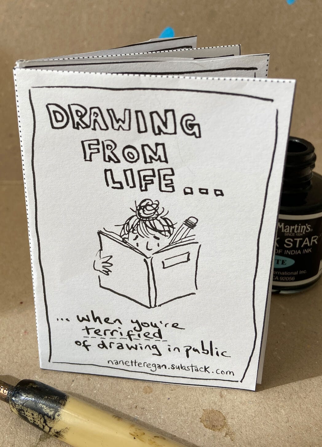 Photo of a handmade zine by Nanette Regan. The title reads Drawing from Life when you're terrified of drawing in public. A pot of ink and dip pen are near the zine