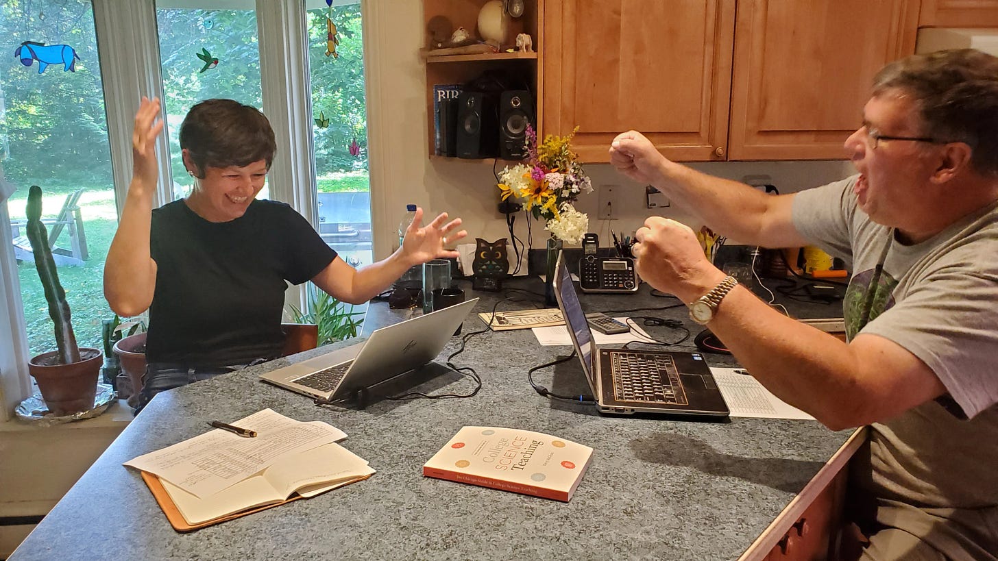 Two people sit opposite each other at a kitchen counter. They both have laptops open and papers out in front of them. They are both smiling with their hands raised in the air. Two people sit opposite each other at a kitchen counter. They both have laptops open and papers out in front of them. They are both smiling with their hands raised in the air.