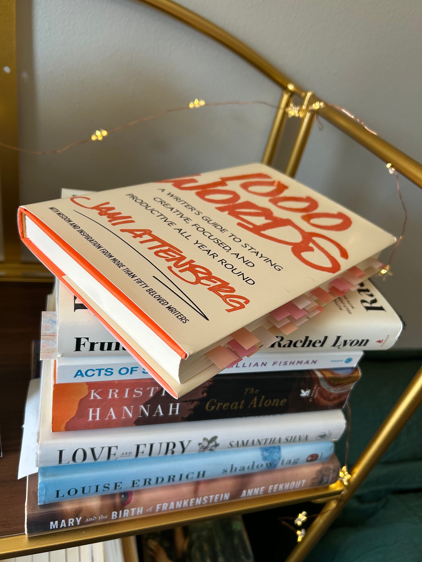 A hardcover copy of 1000 Words with dozens of pink and orange tabs on top of a partial TBR pile.