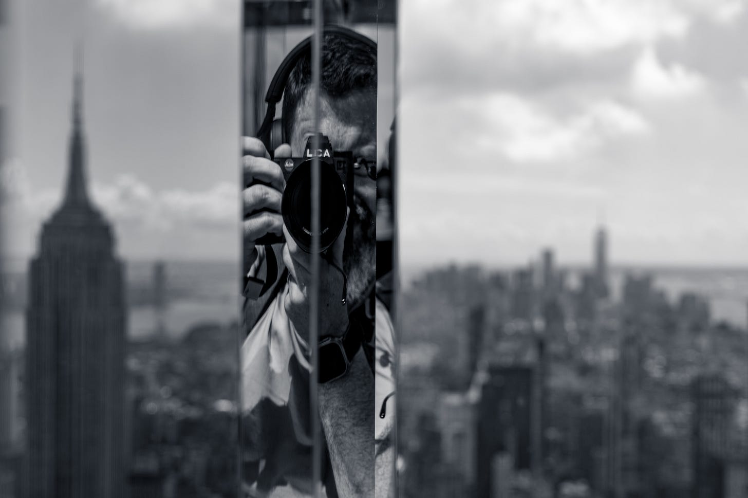 The photographer Paul O'Malley reflected in a narrow mirrored column with a Leica raised to his eye, the Empire State Building and the Midtown skyline visible either side, at Summit One Vanderbilt, New York, in black and white.