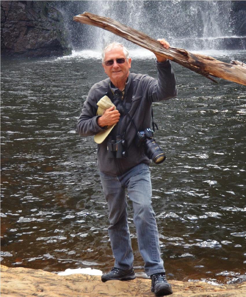 Man standing next to a body of water and waterfall, holding up a large piece of wood