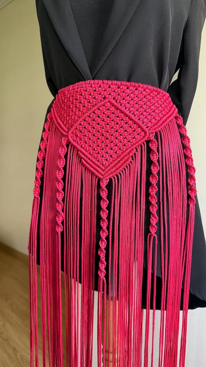 May include: A vibrant fuchsia macrame belt with a geometric diamond pattern and long fringe. The belt is made of knotted cord and features decorative spiral details. This handmade accessory adds a bohemian flair to any outfit.