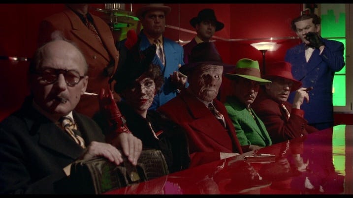 A group of gangsters in vividly colored suits sit around a red-lit table, each with distinct makeup and stylized outfits—highlighting the exaggerated, comic-inspired character design.