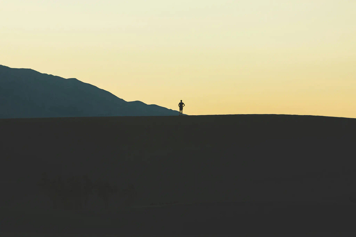 Silhouette of a man standing with arms on his waist on a valley top, looking into the distance. The sunset sky with a hint of yellow and valleys in the background adds a sense of vastness and contemplation.