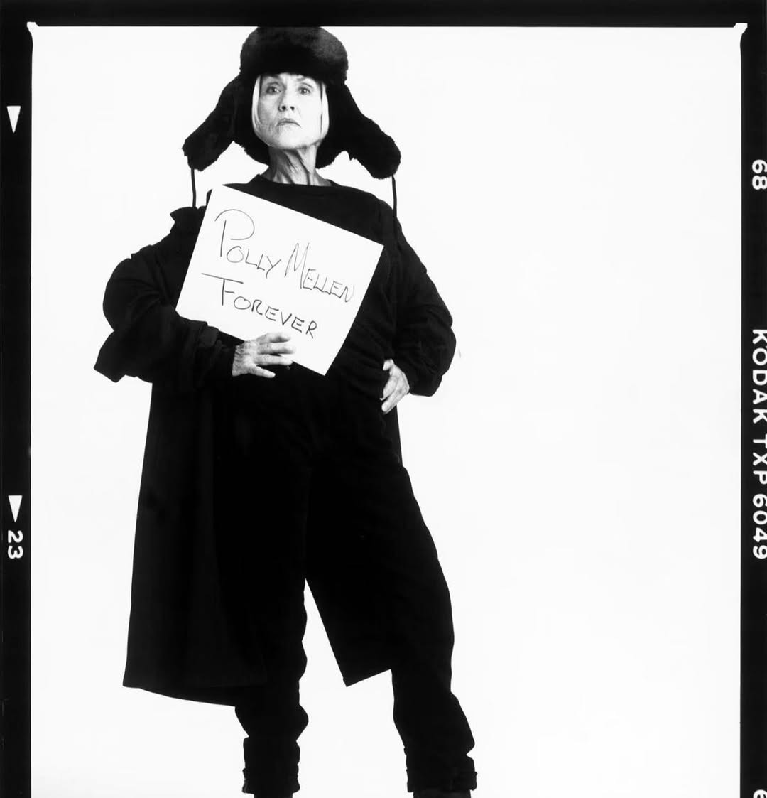 A black and white photo depicts a woman wearing a black fur hat, coat and pants while holding a sign that reads "Polly Mellen Forever"