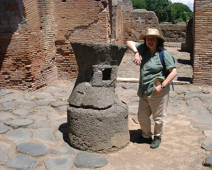 Two Roman grindstones, one form Ostia, one from Pompeii. Nearly the height of the author as the first picture shows with her standing next to a grindstone. Several brick walls stretch away in the left hand picture's background, a fine arched brick oven in the right hand picture.. 