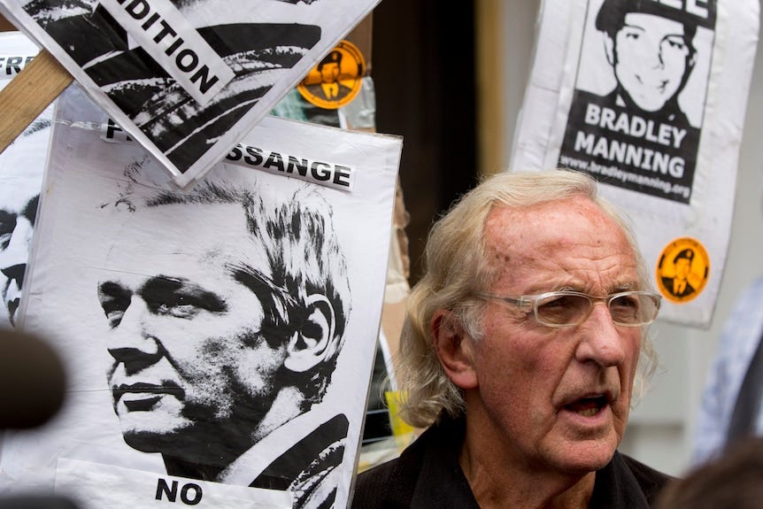 Un vieil homme aux cheveux blonds se tient au milieu dâune mer dâaffiches en noir et blanc de Julian Assange et Bradley Manning. Un vieil homme aux cheveux blonds se tient au milieu dâune mer dâaffiches en noir et blanc de Julian Assange et Bradley Manning.