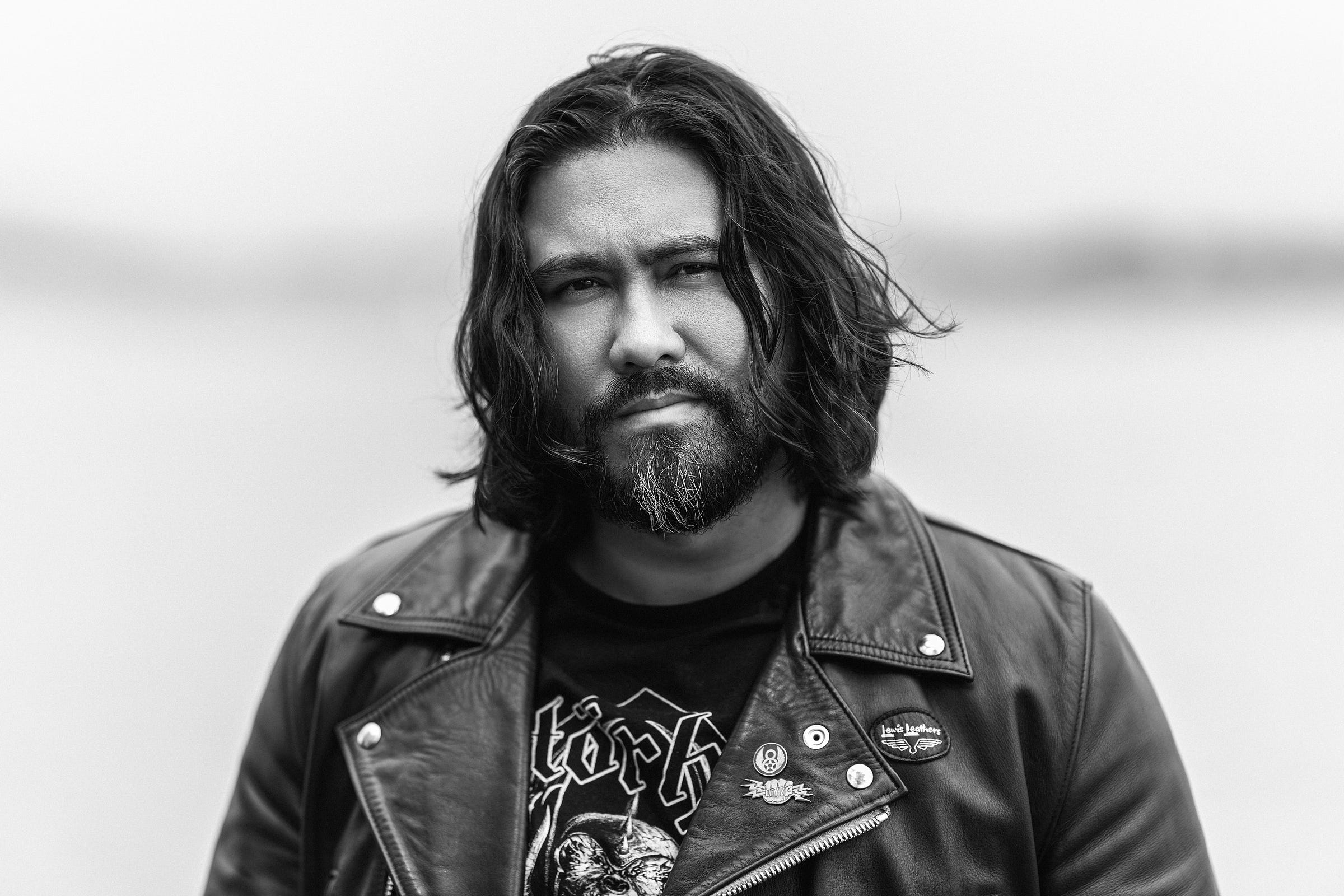 Black and white photograph depicts a mixed race male with long hair and a beard wearing a leather jacket. Black and white photograph depicts a mixed race male with long hair and a beard wearing a leather jacket.