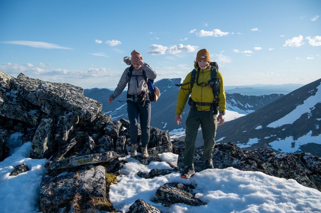 Two hikers cross a rocky, snow-patched ridgetop, one raising an arm across their face against the wind, distant peaks under a partly cloudy sky behind.
