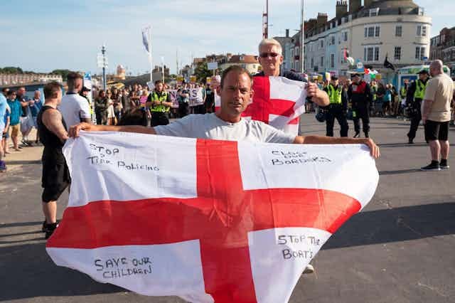 Two men wave English flags in front of a crowd. Writing on the flags reads 'stop two tier policing', 'close our borders', 'save our children' and 'stop the boats'