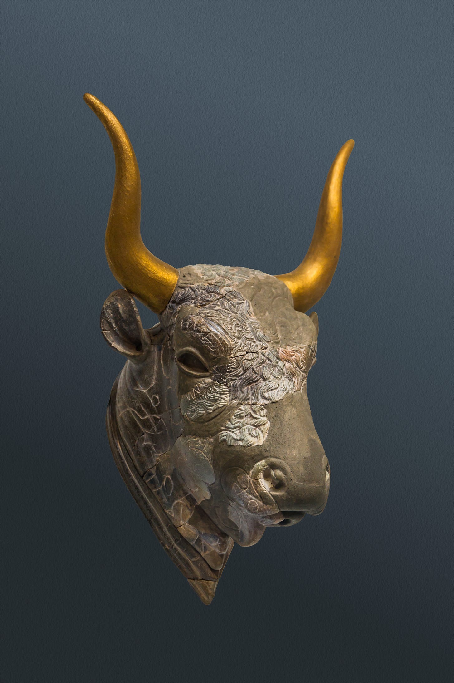 Carved dark grey stone vessel in the shape of a bull's head, with gilded horns. The vessel has multiple cracks and has been reconstructed. The eye sockets are empty, as if stones had been set in them which have since been lost. The animal's head is covered in textured curly hair carved into the stone. A small hole in the lower lip is visible, through which liquid would have poured when in use.