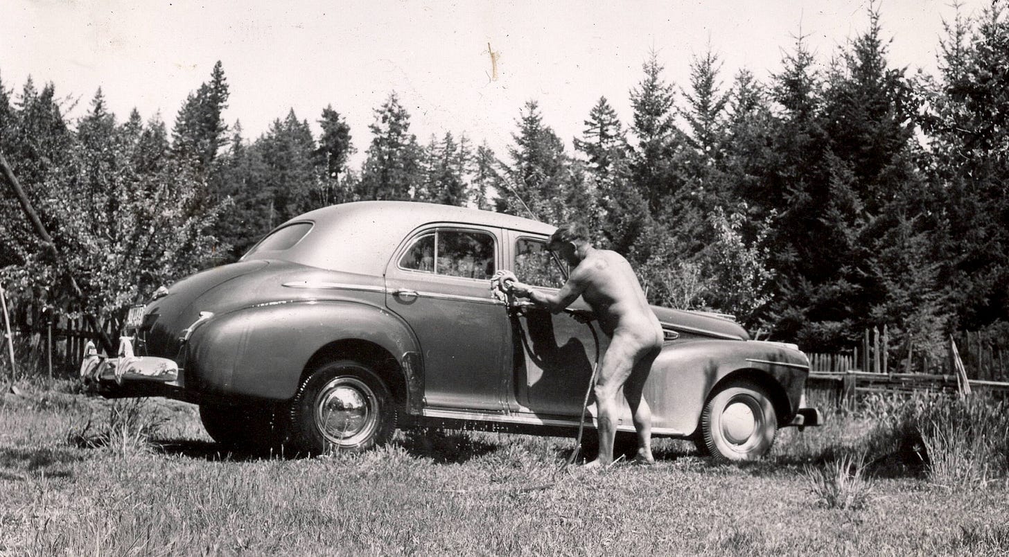 Black-and-white photo of a nude man bending to wash the side of a vintage sedan with a rag, a garden hose coiled at his feet, in a grassy clearing ringed by tall pines. Black-and-white photo of a nude man bending to wash the side of a vintage sedan with a rag, a garden hose coiled at his feet, in a grassy clearing ringed by tall pines.