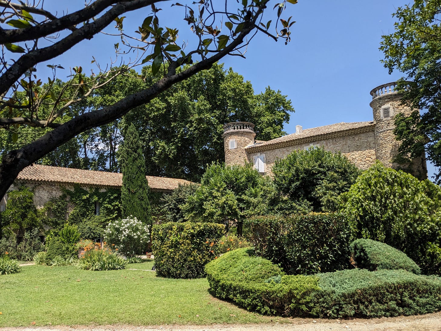 A small French chateau in sand-colored stone fronted by green shrubbery under a blue sky. A small French chateau in sand-colored stone fronted by green shrubbery under a blue sky.